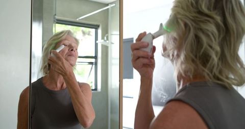 Senior woman using handheld led skincare device at bathroom mirror for anti-aging routine