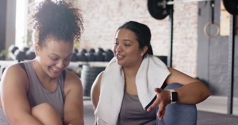 Women chatting and laughing after workout in urban gym, resting with towel and weights