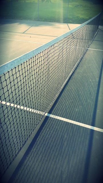 Ideal for illustrating tennis and outdoor sports activities, this photo can be used on sports blogs, in exercise pamphlets, or recreational event posters. It captures an empty tennis court with a net casting a shadow, suggesting a calm, serene athletic environment which can signify preparation or anticipation.