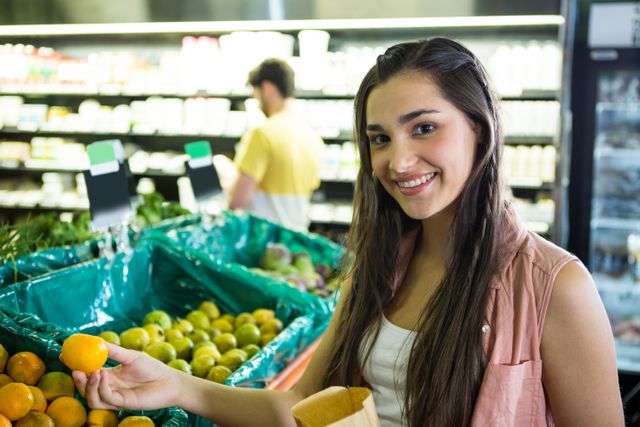 Young woman smiling while selecting fresh fruit in a supermarket. Ideal for use in advertisements for grocery stores, healthy eating campaigns, lifestyle blogs, and retail promotions. Highlights the importance of fresh produce and a healthy lifestyle.