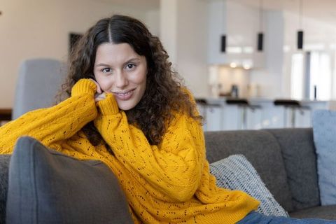 Cozy living room portrait smiling woman wearing mustard-yellow sweater on gray sofa