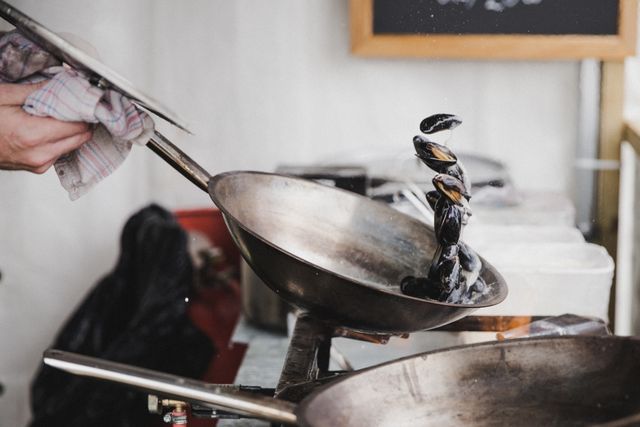 This image depicts a chef preparing mussels in a rustic kitchen setting. The mussels are being tossed in a frying pan, showcasing the chef's professional skills and precision in food preparation. This image can be used for culinary magazines, restaurant advertisements, cooking blogs, or any content related to gourmet seafood dishes and professional cooking techniques.