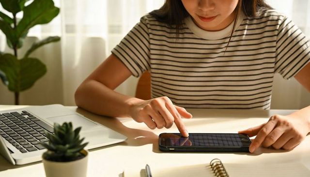 Female tapping smartphone while working at home desk with laptop, notebook and succulent plant