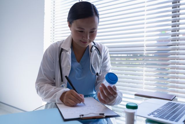 Female doctor in clinic writing a prescription on a clipboard while holding a medicine bottle. Ideal for use in healthcare, medical, and pharmaceutical contexts, showcasing professional medical care and patient treatment.