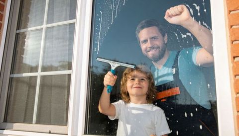 Father and child cleaning sunlit window together with blue squeegee, home chore bonding
