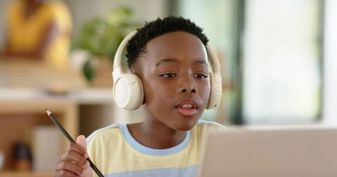 African american boy wearing headphones and holding pencil studying on laptop
