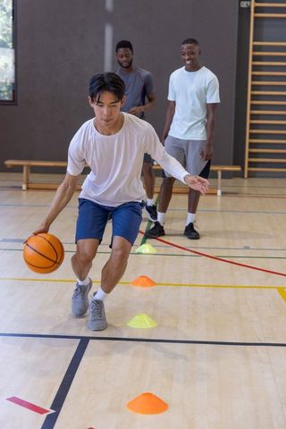 Young men practicing basketball dribbling drill through cones in indoor gym training