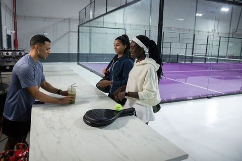 Diverse female padel players receiving green smoothie at club reception near purple court