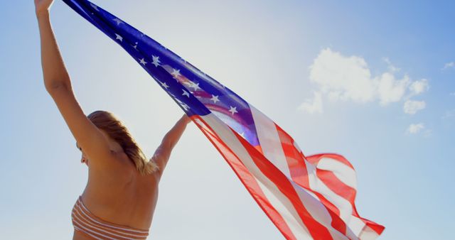 A young Caucasian woman holds the American flag high against a clear sky, with copy space. Her gesture could symbolize patriotism, freedom, or celebration of national events.