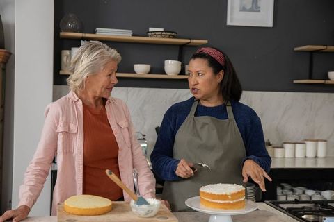 Mature women frosting layered cake together in cozy home kitchen, baking and sharing