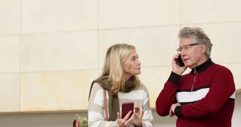 Mature couple using smartphones in modern lobby, woman checking red phone, casual knitwear
