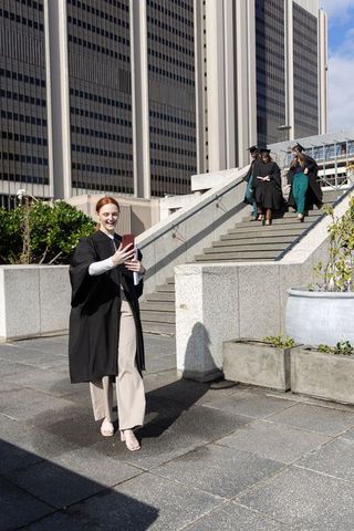Graduate taking selfie in gown on urban plaza while classmates descend stairs in mortarboards