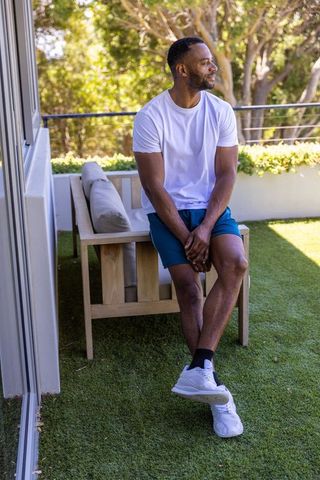 African american man sitting on sunlit terrace in white tee and blue shorts, relaxed