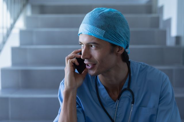 Male surgeon in blue scrubs with a stethoscope around his neck, sitting on hospital stairs and talking on a mobile phone. Ideal for use in healthcare, medical communication, and hospital-related content.