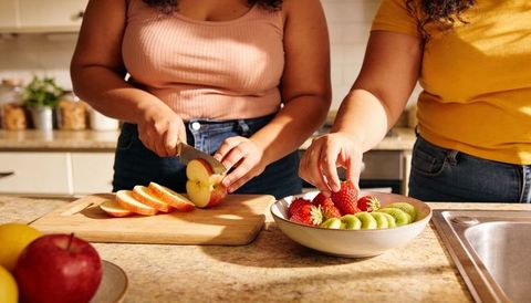 Women slicing apples and arranging strawberries and kiwi on counter preparing fresh fruit bowl