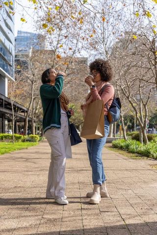 Diverse friends sipping coffee and carrying shopping bags in urban autumn park