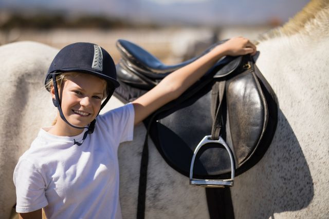 Young boy wearing helmet standing next to white horse, smiling and holding saddle. Ideal for themes related to equestrian activities, children's hobbies, outdoor leisure, and animal companionship.