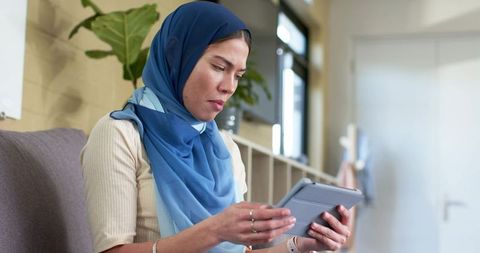 Indian woman wearing blue hijab reading tablet in minimalist living room