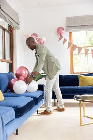 Man placing rose gold white balloons on navy velvet sofa preparing birthday decor