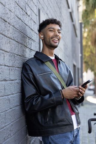 Young man leaning on urban brick wall smiling while holding smartphone with earbuds casual style