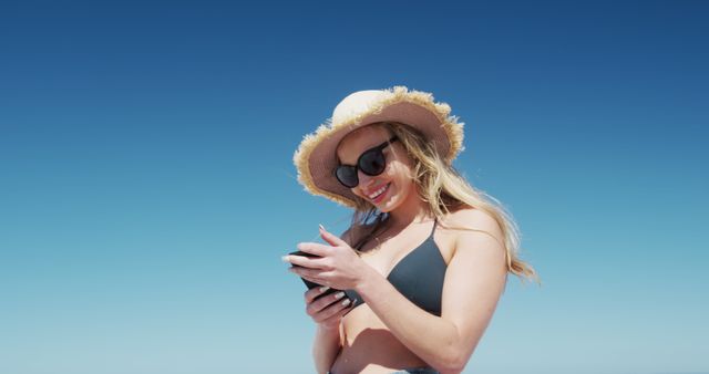 This image features a woman wearing a black bikini and sun hat, smiling as she texts on her smartphone. She is standing outdoors under a clear blue sky. Ideal for use in summer vacation promotions, beachwear advertisements, travel blogs, and lifestyle magazines.