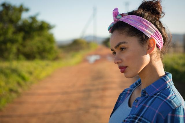 Woman standing on a dirt road in the countryside, looking thoughtful. She is wearing a plaid shirt and a headscarf, with a serene expression. This image can be used for themes related to contemplation, rural life, nature, and casual fashion.