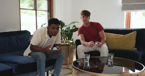 Two Friends Playing Video Game on Living Room Sofas Holding White Controllers, Casual Fun