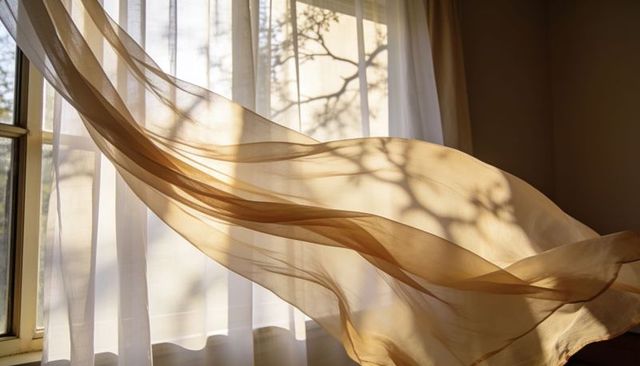 Sunlit sheer curtains billowing with flowing beige drape casting tree-branch shadows