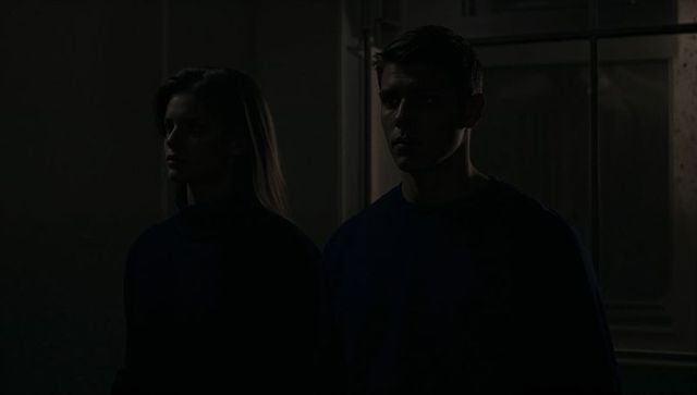 Moody low-key portrait of couple standing in shadow by glass door wearing dark sweaters