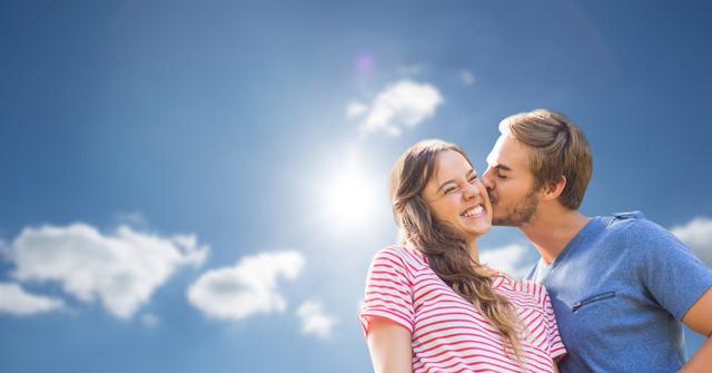 This image shows a young couple sharing an affectionate moment. The woman is smiling while the man is kissing her on the cheek. They are standing outside under a clear blue sky with a few clouds, and the sunlight enhances the warm and joyful atmosphere. This image can be used for advertisements, blog posts, or social media content dealing with love, relationships, happiness, or outdoor activities.
