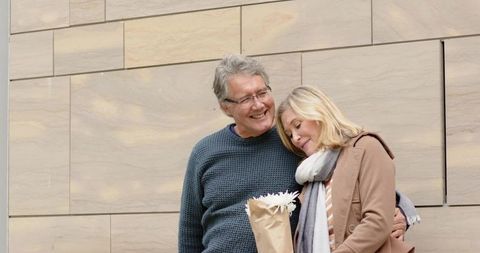 Mature couple sharing tender moment with bouquet against pale stone wall