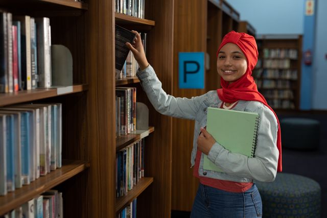 Front view of an Asian female student wearing a red hijab studying in a library selecting a book from a bookshelf, turning and smiling to camera.