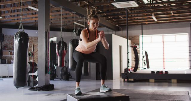Young woman focused on performing a box jump exercise in an industrial-style gym. She is using her fitness routine to build strength and agility. Image ideal for use in fitness blogs, training programs, workout tutorials, and health and wellness promotions.