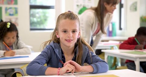 Young Girl Smiling in Classroom with Classmates and Teacher Assisting
