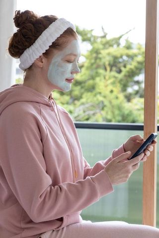 Woman relaxing on balcony with facial mask and headband using smartphone in morning light