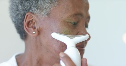 Senior african woman using facial massager along cheek under nose closeup skincare