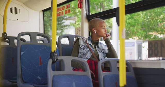 A woman, casually dressed in a denim vest and green sleeves, is sitting alone in a bus and looking out of the window with a thoughtful expression. She is listening to something with her earbuds, enhancing the sense of solitude and reflection. The sun is shining, casting light on the dynamic backdrop outside the bus. Perfect for themes of public transportation, solo travel, relaxed moments, and urban life.