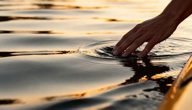 Right hand skimming golden water at sunset creating ripples, droplets and calm reflections