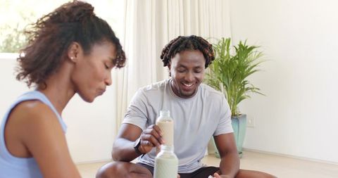 African american couple sharing smoothies sitting on living room floor, minimalist bright home