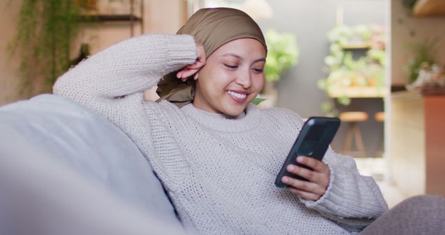 Image of happy biracial woman in hijab using smartphone sitting in living room at home. Happiness, relaxation, communication, inclusivity and domestic life.