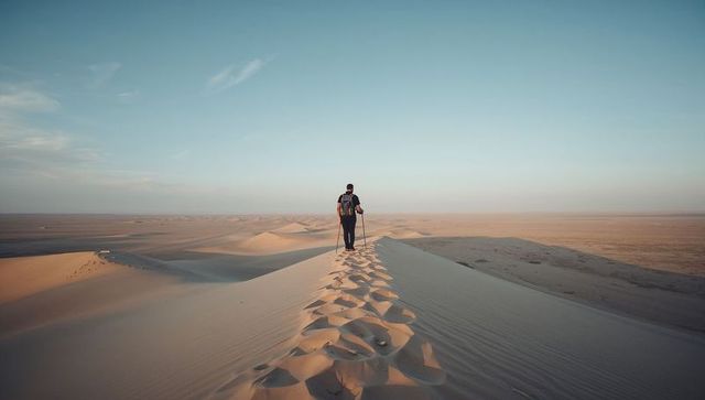 Walking dune crest carrying backpack and trekking pole overlooking vast desert horizon