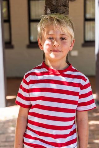 Young blond boy wearing red white striped shirt standing by tree smiling naturally