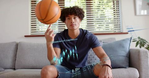 Young man spinning basketball on finger at home with holographic financial data overlay