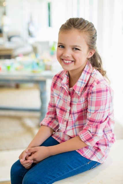 Young girl wearing a pink plaid shirt and jeans, sitting indoors with a cheerful smile. Ideal for use in family-oriented content, lifestyle blogs, advertisements for children's clothing, or educational materials.