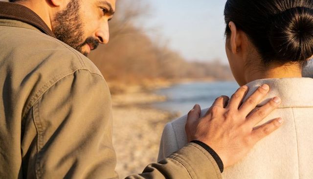 Man comforting woman on riverbank at golden hour, hand on shoulder, calm moment