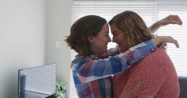 This lovely image displays two women embracing each other while smiling intimately, reflecting a happy and affectionate moment at home. The sunlight filtering through closed blinds adds a cozy atmosphere, enhancing the sense of warmth and love between them. Perfect for use in content related to love, LGBTQ+, relationships, togetherness, and domestic bliss.