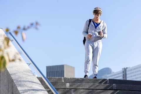 Young man standing on urban steps holding smartphone and coffee wearing gray tracksuit