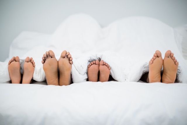 Family feet peeking from under a white blanket in bed, showcasing togetherness and comfort. Ideal for use in articles or advertisements related to family life, parenting, home comfort, and relaxation. Perfect for illustrating concepts of bonding, morning routines, and cozy home environments.