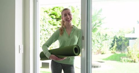 Smiling woman entering backyard holding green yoga mat for home workout