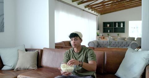 African american man sitting on leather sofa reaching into popcorn bowl, cozy living room